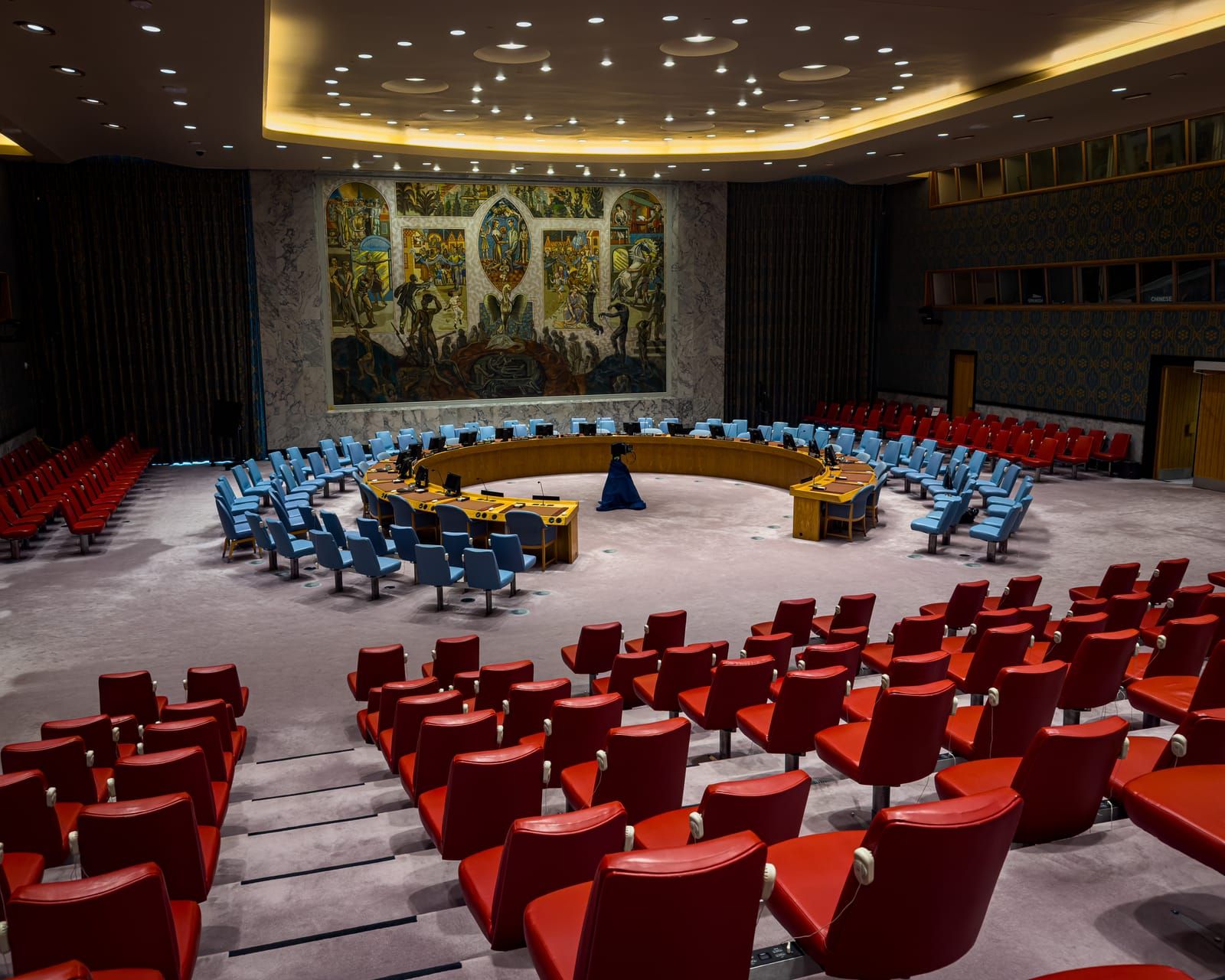 The empty UN Security Council chamber in New York: a horseshoe table ringed by blue chairs beneath a large mural, with rows of red gallery seats in the foreground