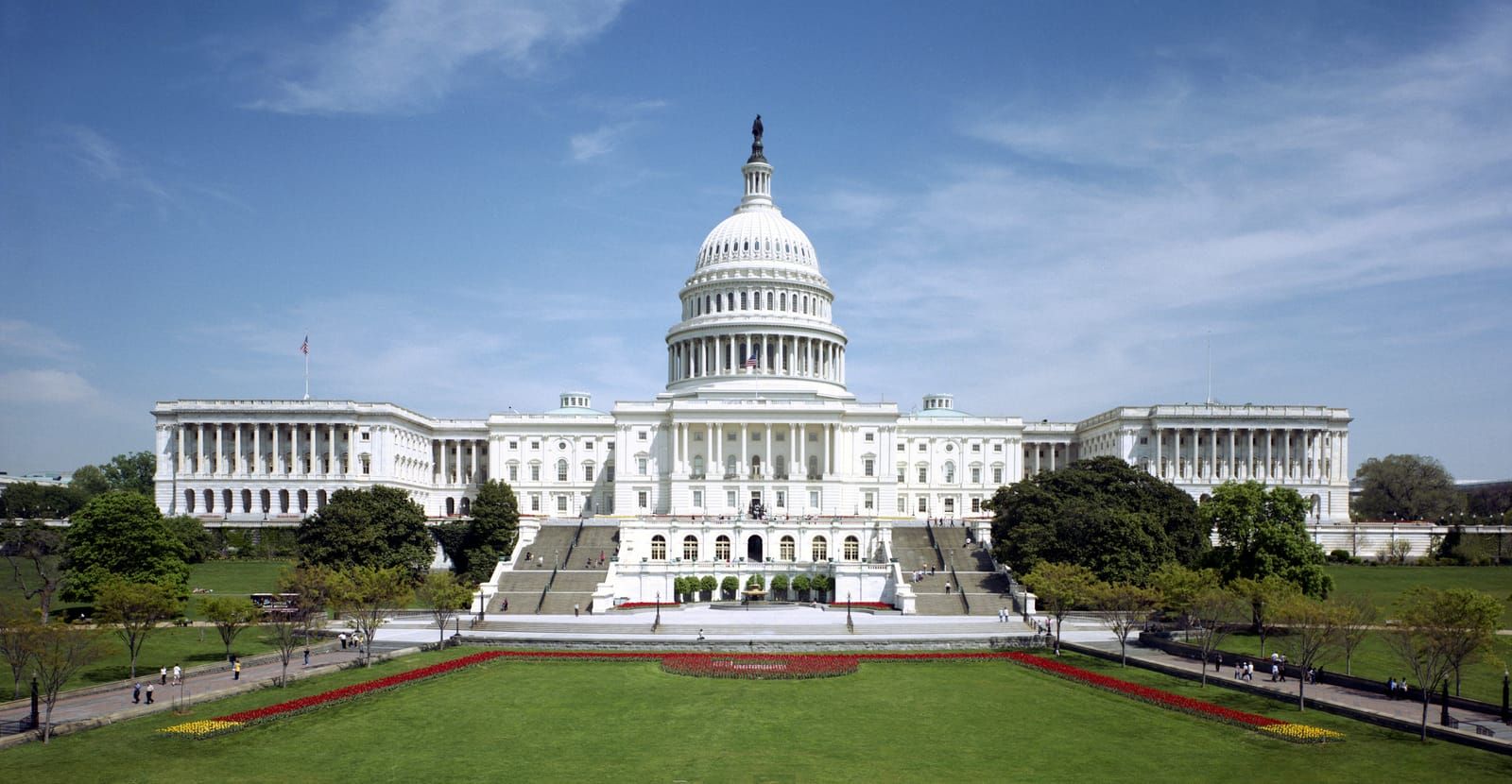 The west front of the United States Capitol in Washington under a blue sky, with a lawn and flower beds in front
